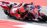 Red MotoGP rider in red leathers leaning into a corner on a sponsor-covered race bike.