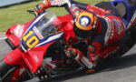 Racing motorcycle rider in red-blue Honda livery leans into a curve, wearing a colorful AGV helmet and number 10 plate on the bike