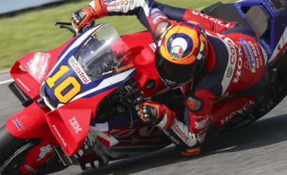 Racing motorcycle rider in red-blue Honda livery leans into a curve, wearing a colorful AGV helmet and number 10 plate on the bike