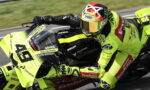 Motorsport rider on a neon yellow Ducati race bike leaning into a turn at speed, wearing matching neon leathers and helmet.