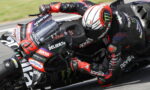 Rider in red and black leathers leaning into a turn on an Aprilia race bike with sponsor logos visible on the fairing.