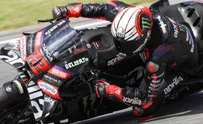 Rider in red and black leathers leaning into a turn on an Aprilia race bike with sponsor logos visible on the fairing.