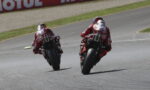 Two motorcycle racers in red racing suits lean into a left-hand bend on a paved race track, with a red Motul banner in the background.