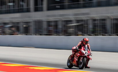 Motorcycle racer in a red racing suit leaning into a turn on a race track. The rider and bike are in motion with the pit wall in the background.] ,