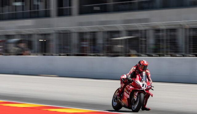 Motorcycle racer in a red racing suit leaning into a turn on a race track. The rider and bike are in motion with the pit wall in the background.] ,