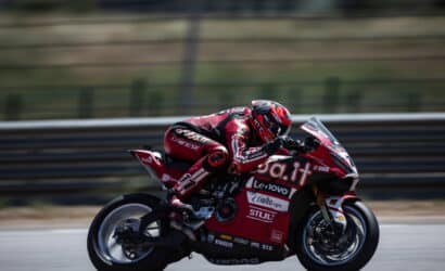Red racing motorcycle and rider in full gear speeding through a turn on a racetrack, sponsor logos visible on the bike's fairing.