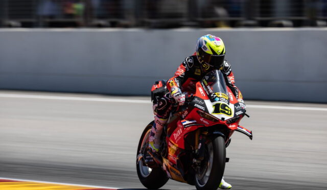 Racer on a red racing bike leaning into a turn on a sunny track, wearing a neon-yellow helmet and red suit.