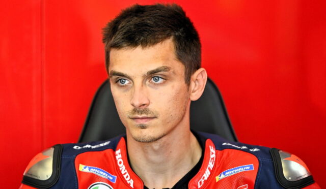 Portrait of a young man in a red and blue motorcycle racing suit against a solid red background, looking off to the side.