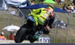 Professional motorcycle racer in a neon-yellow suit riding a Ducati bike, leaning into a sharp turn during a race with spectators in the background
