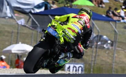 Professional motorcycle racer in a neon-yellow suit riding a Ducati bike, leaning into a sharp turn during a race with spectators in the background