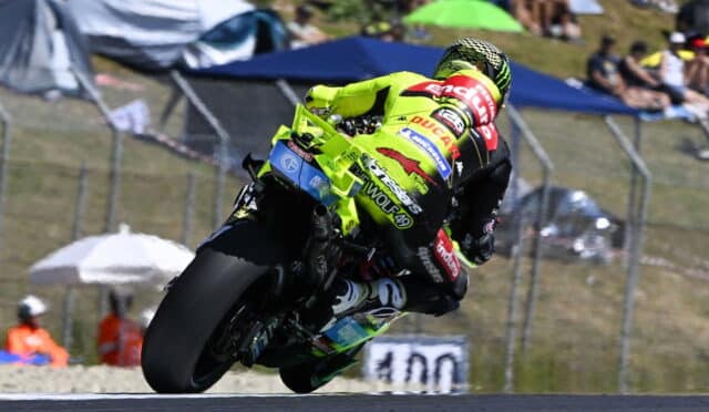 Professional motorcycle racer in a neon-yellow suit riding a Ducati bike, leaning into a sharp turn during a race with spectators in the background