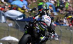 Motorcycle racer in a neon green helmet leaning into a turn on a race track with spectators in the background behind barriers.