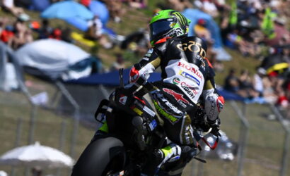 Motorcycle racer in a neon green helmet leaning into a turn on a race track with spectators in the background behind barriers.