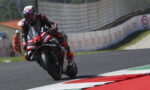 Motorcycle racer in red and black gear leaning into a right-hand turn on a race track with colorful curbs in view.