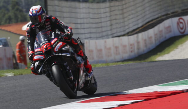 Motorcycle racer in red and black gear leaning into a right-hand turn on a race track with colorful curbs in view.