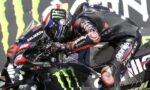 MotoGP rider in black and red gear crouches over a black Monster Energy race bike, leaning into a corner.