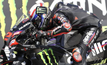 MotoGP rider in black and red gear crouches over a black Monster Energy race bike, leaning into a corner.