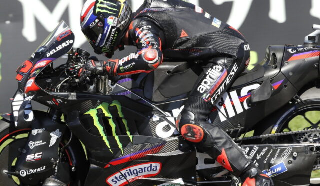 Rider in full race leathers crouches over a black Kawasaki/Aprilia sports bike with Monster Energy logos and sponsor decals during a race. Helmet features Monster branding.