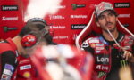 MotoGP rider in a red Lenovo-sponsored racing suit sits in the pit area, holding a drink with a straw and looking off to the side.