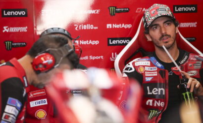 MotoGP rider in a red Lenovo-sponsored racing suit sits in the pit area, holding a drink with a straw and looking off to the side.