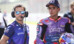 Two motorcycle racers and a team member stand side by side in racing gear; the man on the left wears a blue Primo team shirt, headphones, and reflective sunglasses, while the man on the right wears a purple-red Yamaha/Alpinestars suit and a cap with mirrored sunglasses.