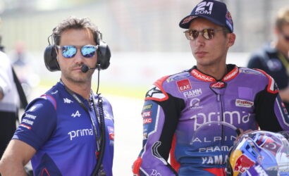 Two motorcycle racers and a team member stand side by side in racing gear; the man on the left wears a blue Primo team shirt, headphones, and reflective sunglasses, while the man on the right wears a purple-red Yamaha/Alpinestars suit and a cap with mirrored sunglasses.