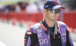 Male motorcycle racer in a purple PRAMAC suit, cap, and sunglasses on a pit lane.