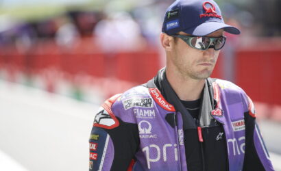 Male motorcycle racer in a purple PRAMAC suit, cap, and sunglasses on a pit lane.