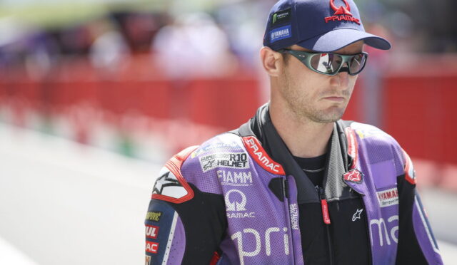 Male motorcycle racer in a purple PRAMAC suit, cap, and sunglasses on a pit lane.