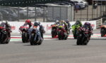 Group of motorcycle racers in a tight pack leaning into a turn on a race track