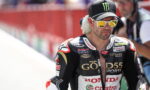 Motorcycle racer in Honda GoD55 racing suit with Castrol branding, wearing a Monster Energy cap and gold mirrored sunglasses in a pit area.