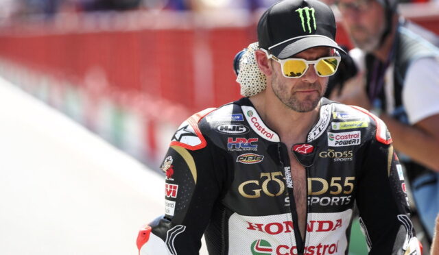Motorcycle racer in Honda GoD55 racing suit with Castrol branding, wearing a Monster Energy cap and gold mirrored sunglasses in a pit area.