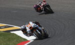 Two motorcycle racers lean into a sharp left turn on a smooth race track, close together as they race side by side.