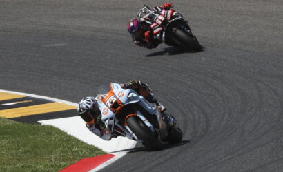 Two motorcycle racers lean into a sharp left turn on a smooth race track, close together as they race side by side.