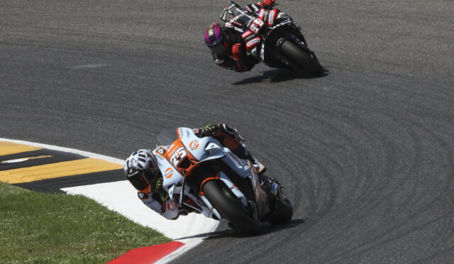 Two motorcycle racers lean into a sharp left turn on a smooth race track, close together as they race side by side.