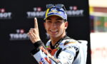 Motorsport racer in a racing suit and cap, smiling and pointing his index finger upward in celebration.