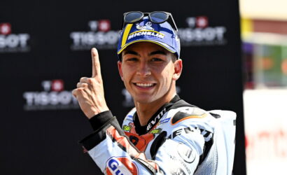 Motorsport racer in a racing suit and cap, smiling and pointing his index finger upward in celebration.