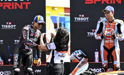 Two motorcyclists on a podium as a staffer hands a medal to the left rider, with sponsor banners behind (TISSOT).