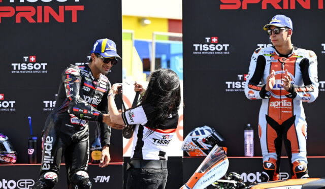 Two motorcyclists on a podium as a staffer hands a medal to the left rider, with sponsor banners behind (TISSOT).