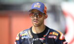 Racer in a blue and orange KTM/Red Bull cap and racing suit wearing large sunglasses, outdoors and focused on something off-camera.
