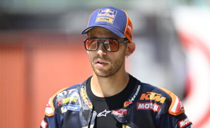 Racer in a blue and orange KTM/Red Bull cap and racing suit wearing large sunglasses, outdoors and focused on something off-camera.
