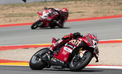 Red sport motorcycle leaning into a sharp turn on a race track, rider in red gear with sponsor decals visible on the bike's fairing.