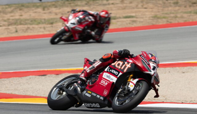 Red sport motorcycle leaning into a sharp turn on a race track, rider in red gear with sponsor decals visible on the bike's fairing.