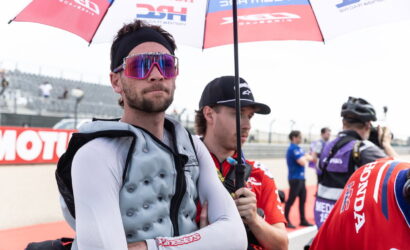 Racer in a gray protective vest and white sleeves stands with arms crossed under a sponsor umbrella at a race track and pit area.
