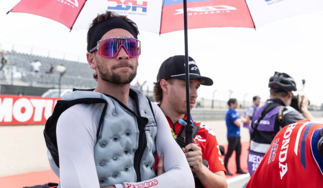 Racer in a gray protective vest and white sleeves stands with arms crossed under a sponsor umbrella at a race track and pit area.