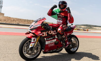 Racing motorcyclist in red suit on a Ducati bike, arms raised in victory on a track with clear blue sky, sponsor logos visible (Aruba.it, Lenovo).