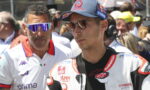 MotoGP rider in white leather racing suit with sponsor logos, wearing brown sunglasses and a blue cap, standing among a crowd and team members.