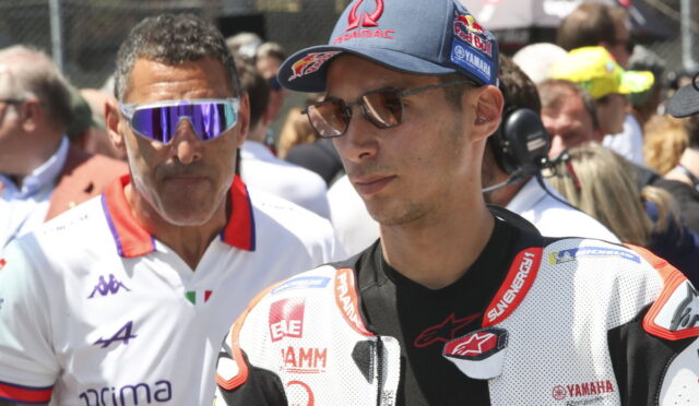 MotoGP rider in white leather racing suit with sponsor logos, wearing brown sunglasses and a blue cap, standing among a crowd and team members.