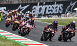 Motorcycle race with riders leaning into a curve on a track, colorful helmets and sponsor logos in view.