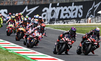 Motorcycle race with riders leaning into a curve on a track, colorful helmets and sponsor logos in view.